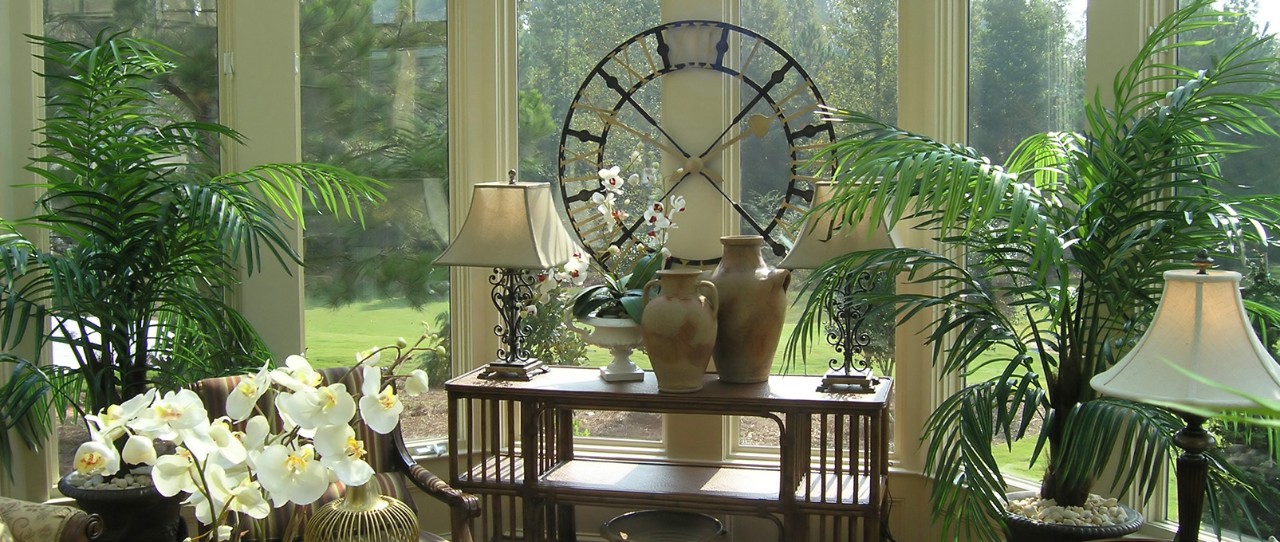 Large sunroom with long side table and lamps along with plant greens everywhere and a large metal clock. 